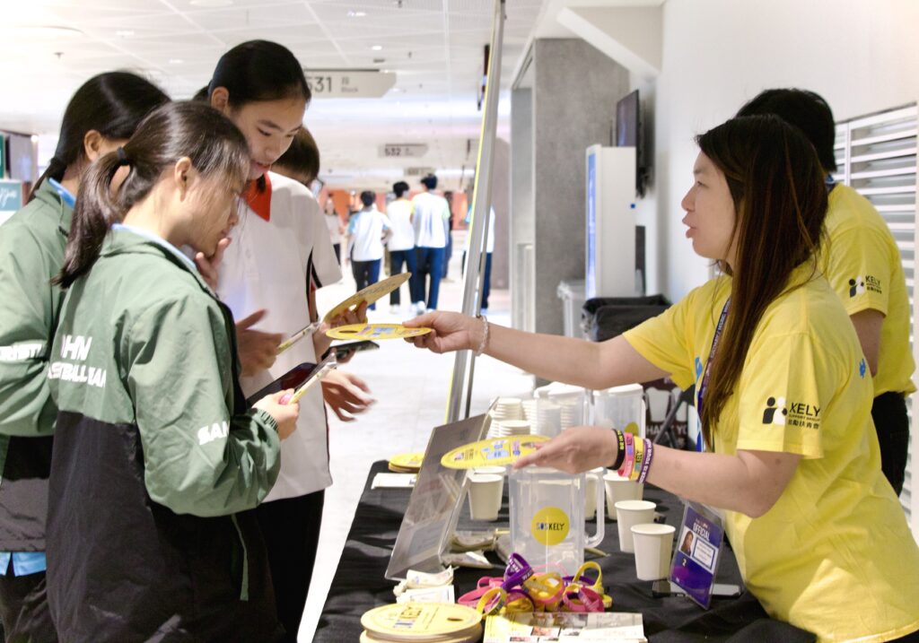 A person in a yellow KELY t-shirt hands a round yellow card to a student in school uniform at an information table.