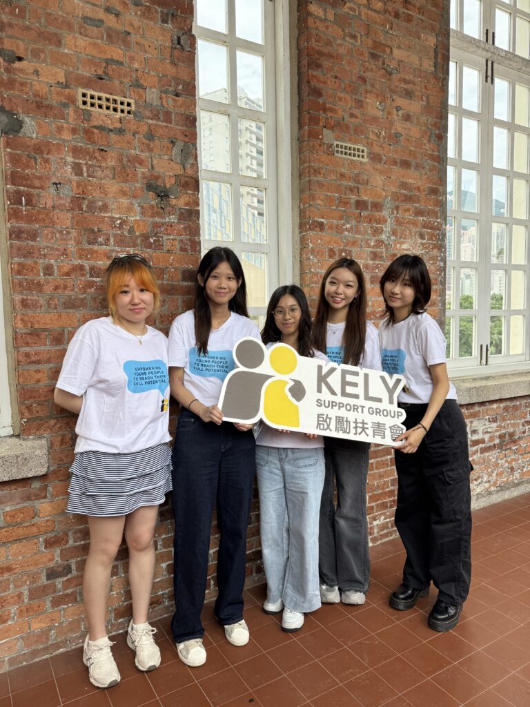 Five young people in white t-shirts stand against a red brick wall, holding a KELY Support Group sign.