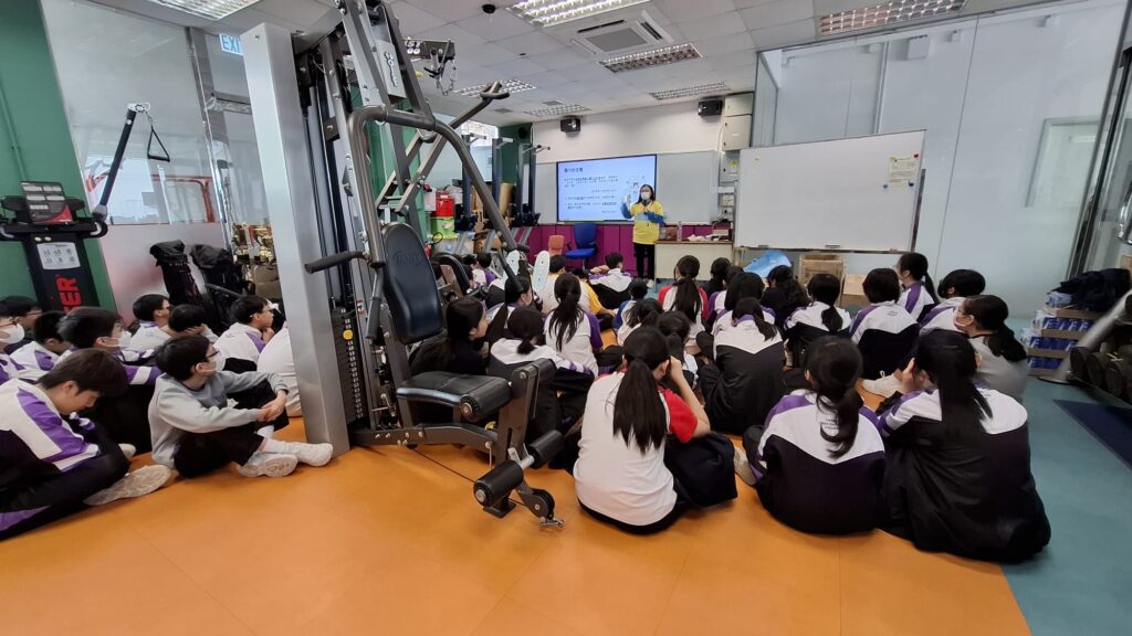 A person in a yellow shirt presents on a screen to students sitting on the floor of a room with exercise equipment.